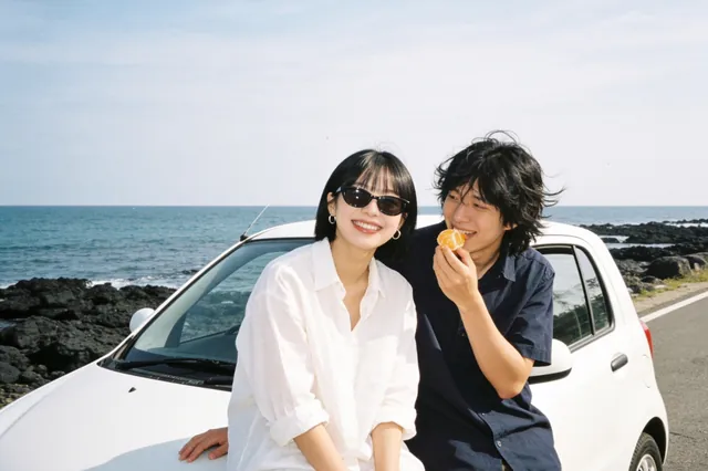 AI 예시 사진: 제주 바닷길에서 차에 기대 귤을 먹으며 웃는 두 사람